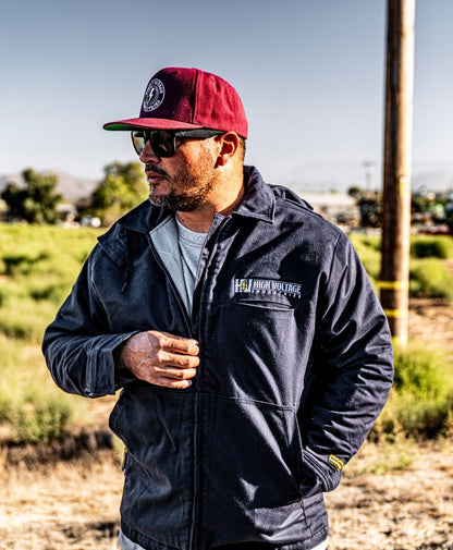 Man wearing a black FR jacket with HVI logo and a maroon bolt trucker outdoors.