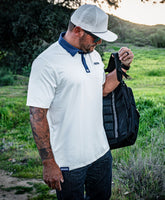 Man in a white polo shirt with a  'High Voltage' logo, holding a black backpack in a natural setting.