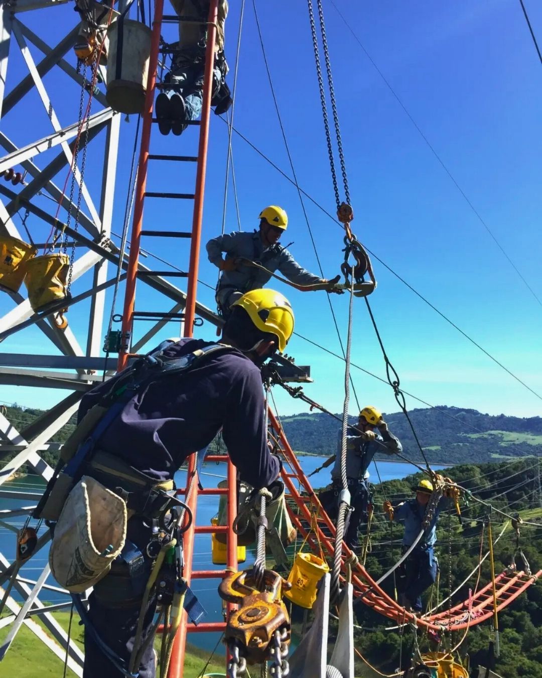 A team of linemen in safety gear working on a transmission tower overlooking a lake.