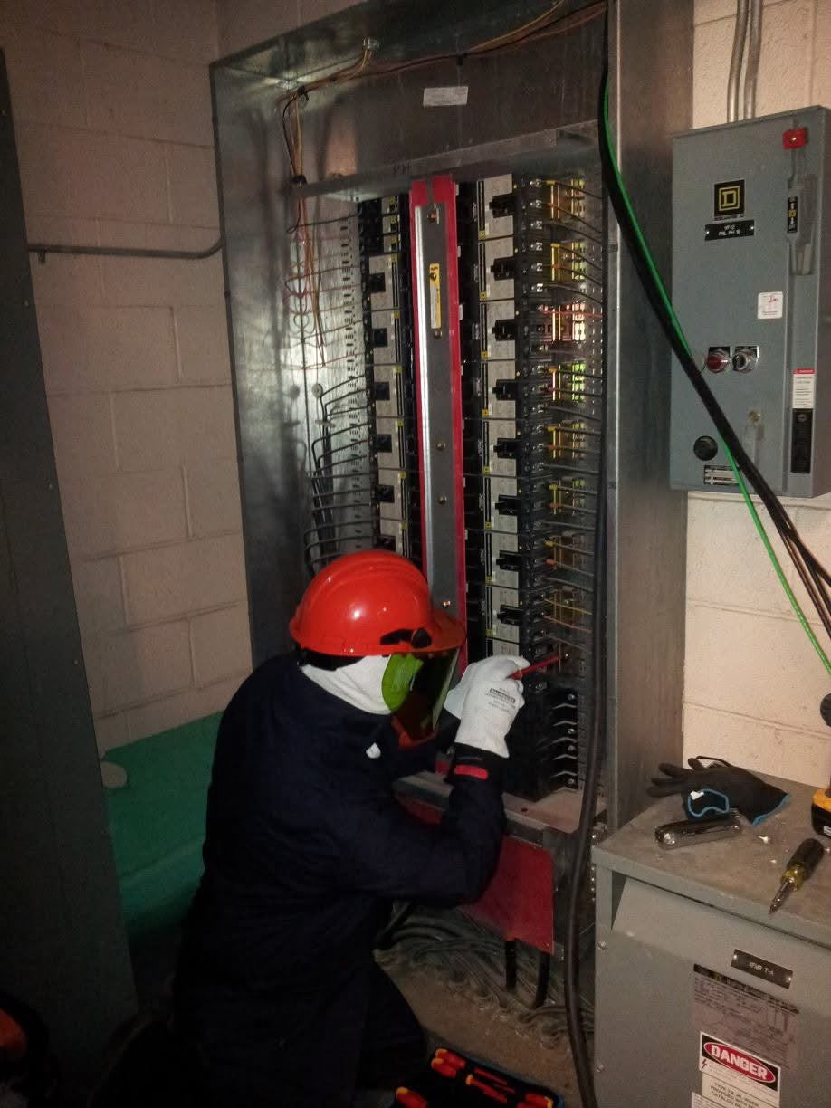 Sergio wearing safety gear inspects industrial electrical panel with insulated tools in a control room.