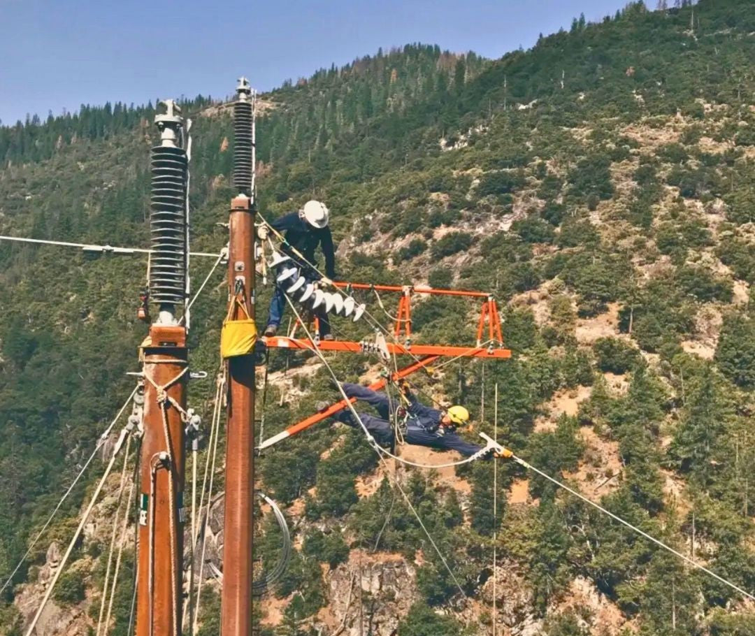 Two linemen working on a utility pole, with one leaning out in a safety harness to service a power line on a steep, forested hill.