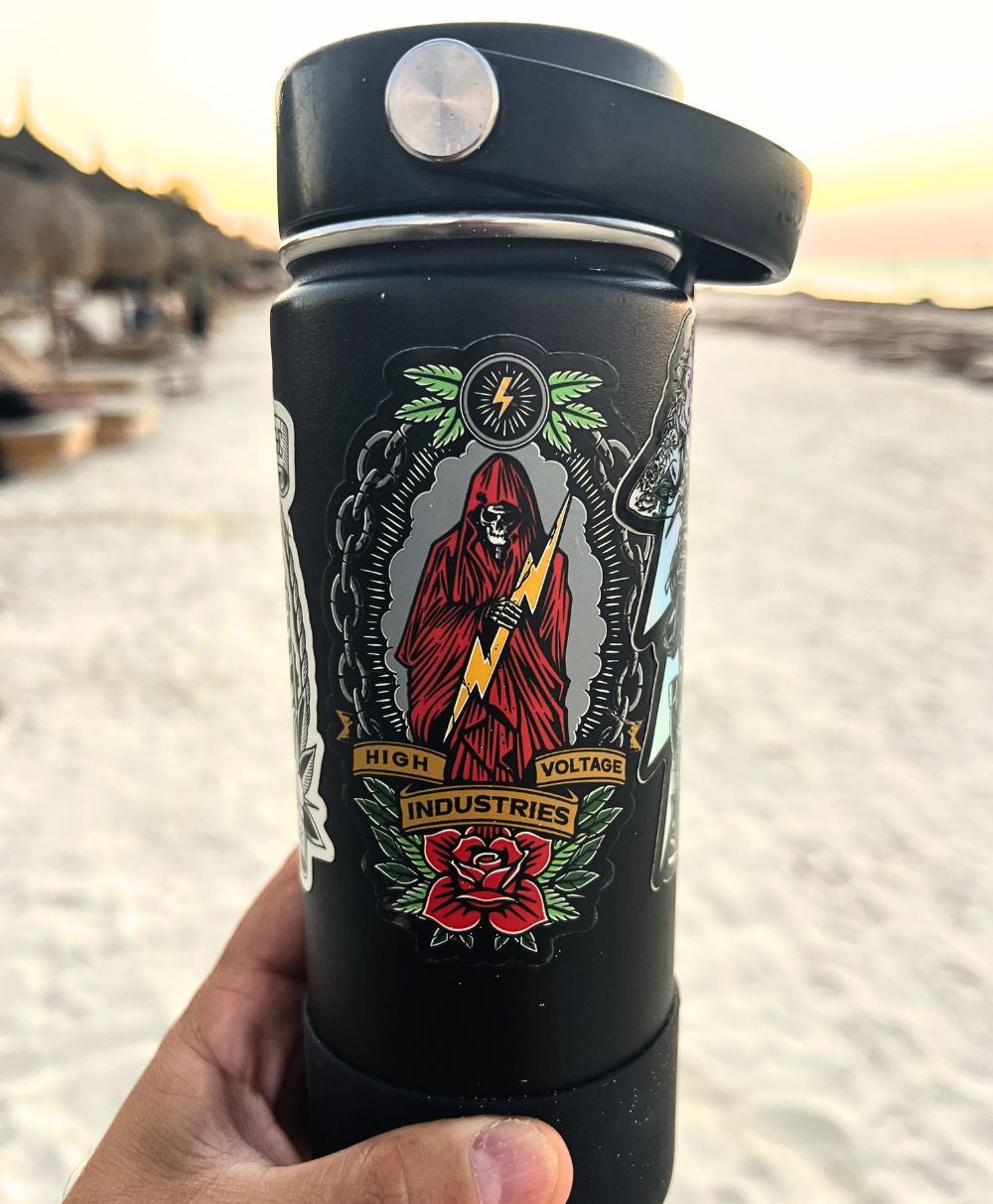 Black insulated mug with a colorful reaper design held in a hand against a blurred outdoor background.