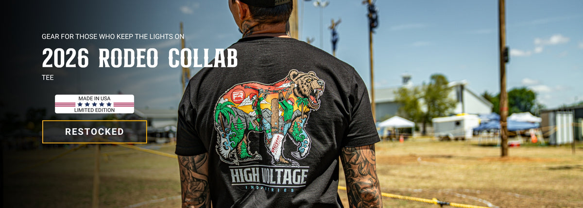 A man viewed from behind wearing a black graphic t-shirt with a colorful bear riding a motorcycle design and the text 'HIGH VOLTAGE' on the back. The setting is an outdoor rodeo arena under a blue sky.